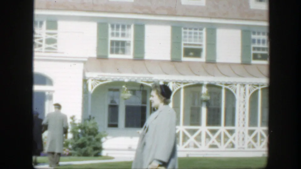 1950 Montreal home-movie frame of adults moving beside garden shrubs and a white porch with green shutters.