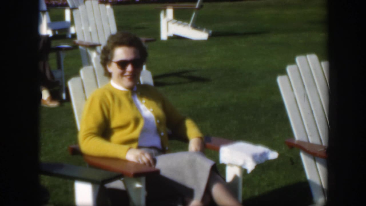 1950 Montreal home-movie frame of an older woman in sunglasses and a yellow cardigan seated on a white Adirondack chair on a lawn.