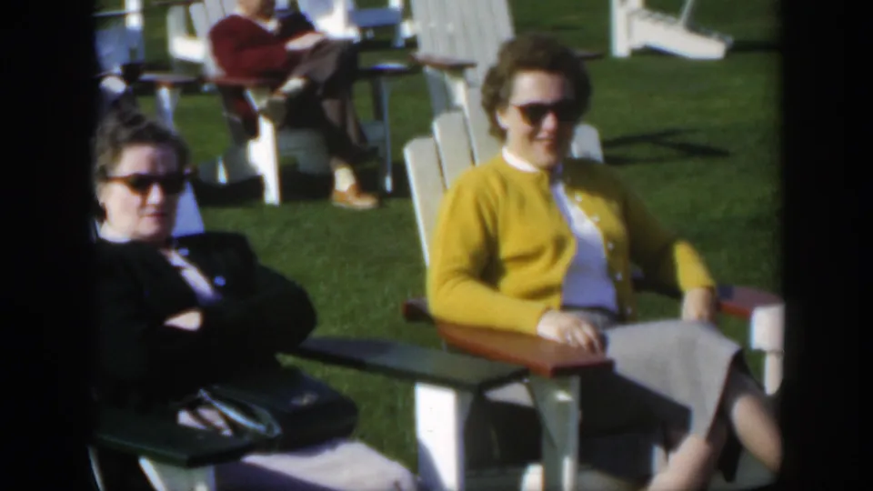 1950 Montreal home-movie frame of two older women in sunglasses seated side by side on white lawn chairs at an outdoor gathering.