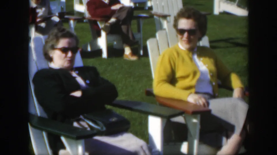1950 Montreal frame showing two older women seated on white Adirondack chairs, one in a yellow cardigan and one in a dark cardigan.