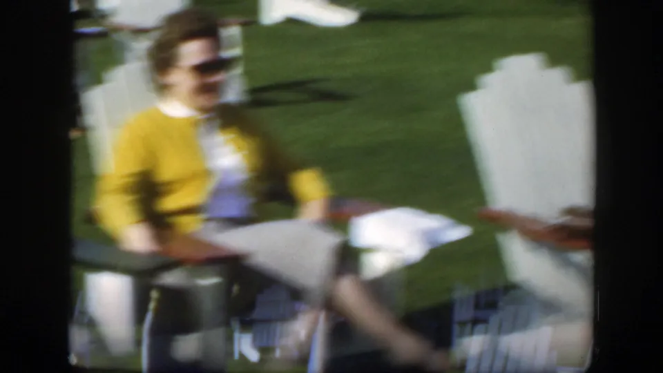 Soft handheld 8mm frame showing the woman in yellow seated on the lawn, with white chairs and grass around her.