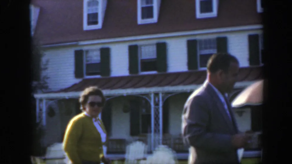 A woman in yellow and a man in a suit appear on a lawn near a white house with shutters in 1950 Montreal footage.