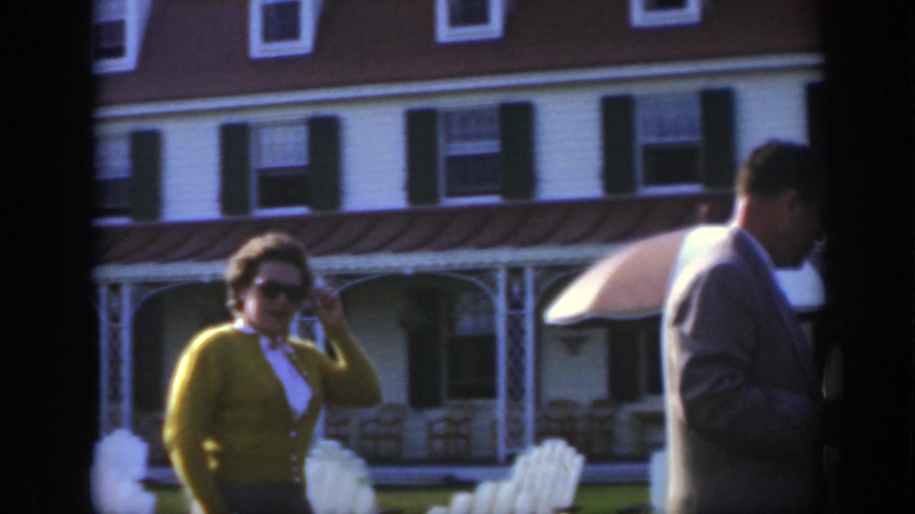 Woman in green cardigan and sunglasses walks past outdoor chairs near a white building.