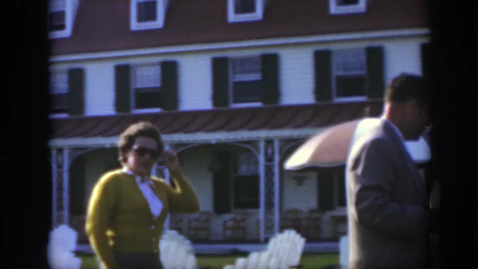 1950 Montreal home-movie frame of a woman in sunglasses and green cardigan walking near white outdoor chairs and a clapboard building.