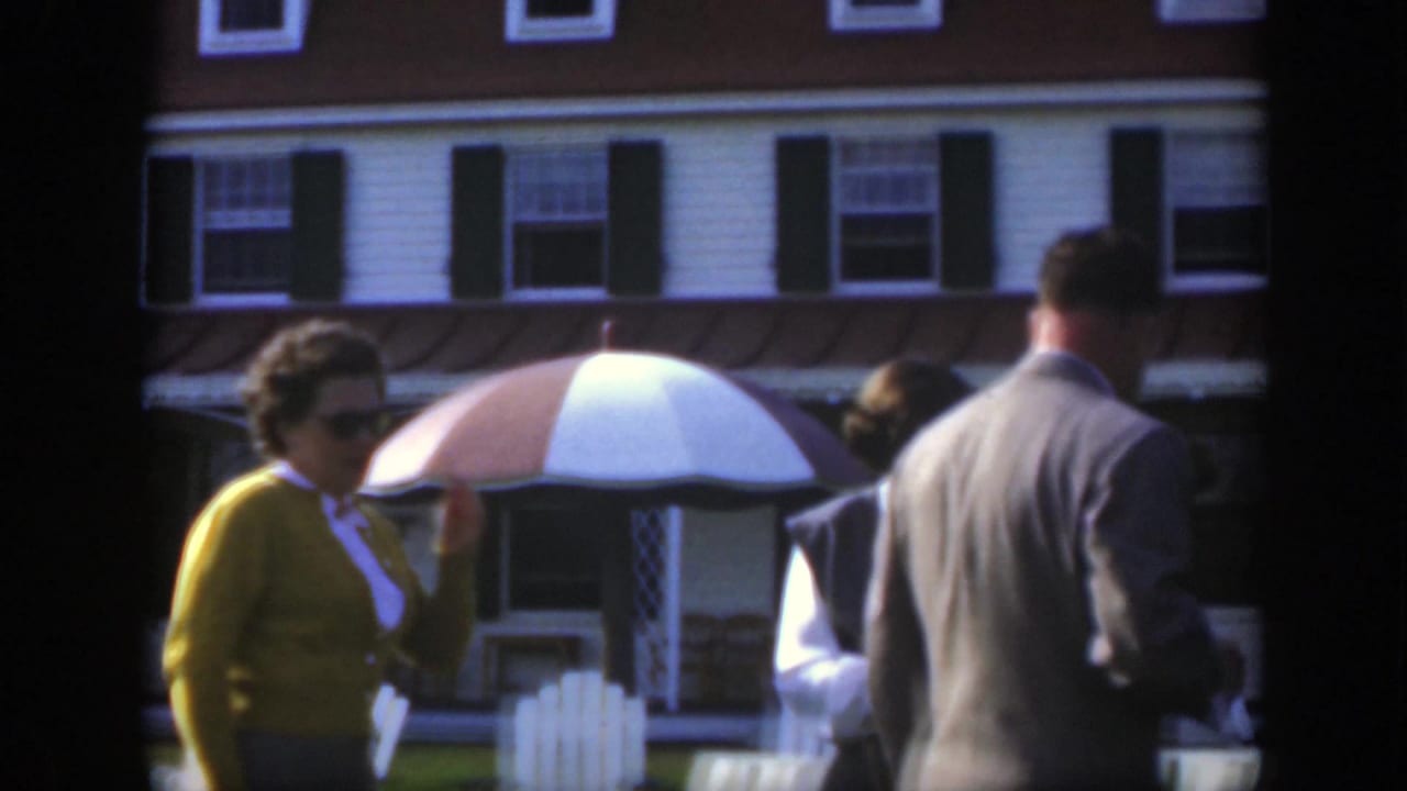 1950 Montreal home-movie frame showing adults outside a white building with dark shutters and a striped patio umbrella.
