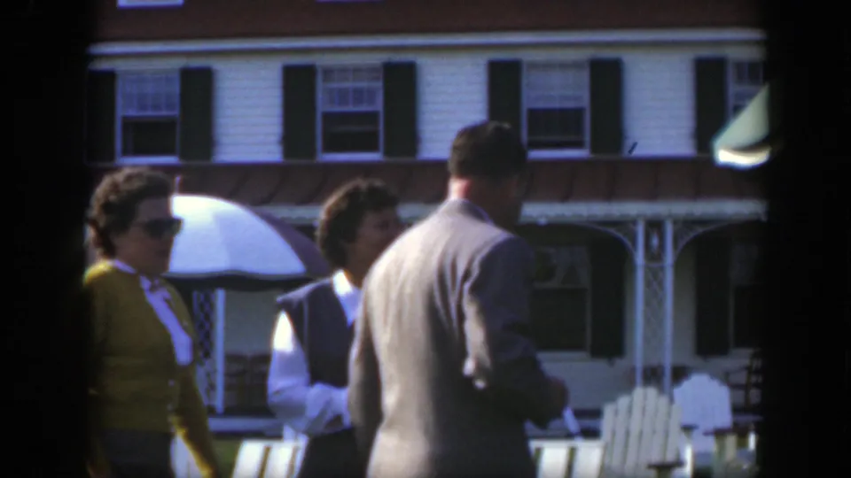 1950 Montreal home-movie frame of adults near white Adirondack chairs, a patio umbrella, and a white clapboard building.