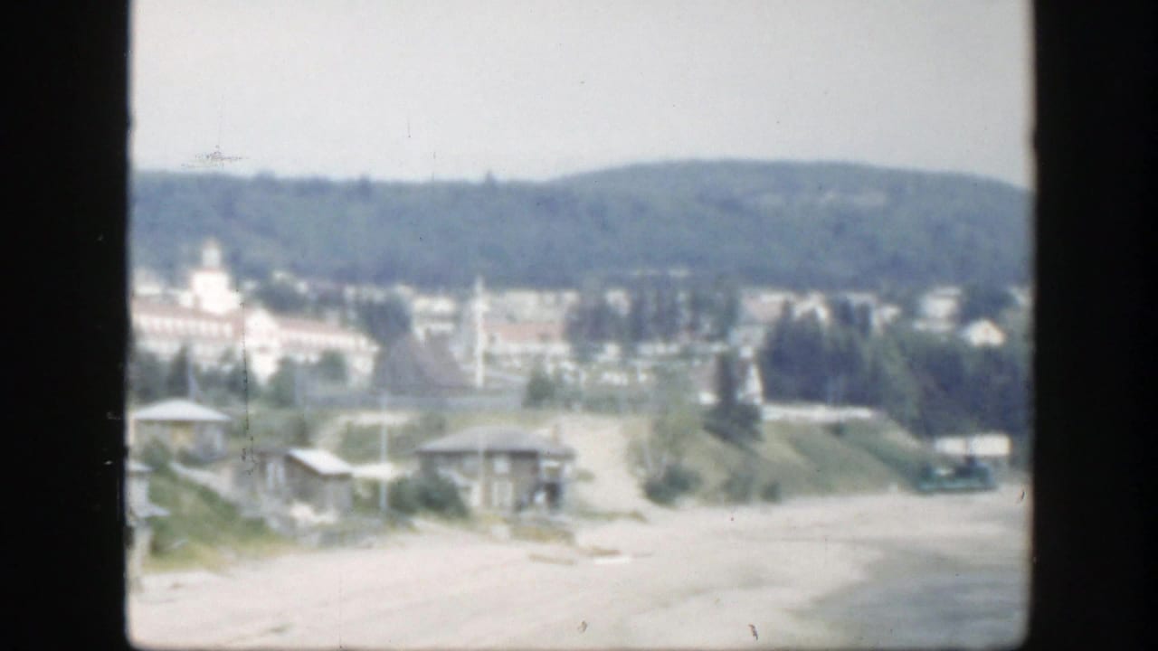 Beachfront road and small buildings with trees, hills, and a distant towered structure.