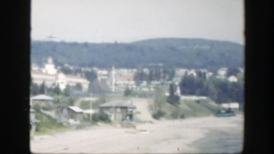 1950 Montreal waterfront view with sandy ground, small roadside buildings, scattered trees, distant hills, and a pale towered building.