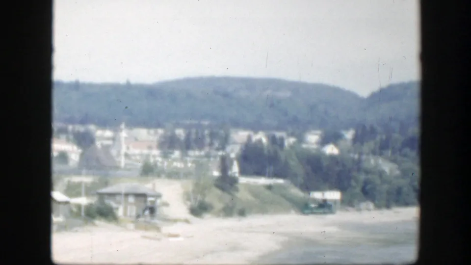 1950 Montreal frame showing a panned shoreline with a small building, grass, trees, hills, and the edge of the beach and water.