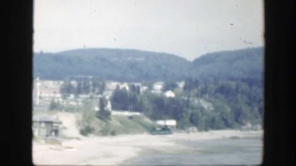1950 Montreal home-movie frame with sandy shoreline, water, green hillside, scattered buildings, and trees in soft focus.
