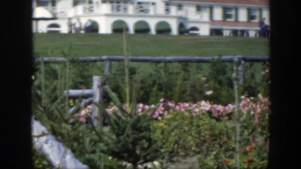 1950 Montreal garden scene with flowers, a low fence, green lawn, and a white arched building as small figures walk nearby.