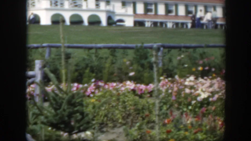 1950 Montreal home-movie frame showing garden flowers, fence, lawn, arched white building, and distant people gathered or walking at right.