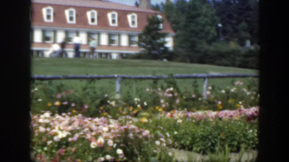 1950 Montreal garden scene with blooming flowers, a low fence, men on the lawn, and trees beside a white red-roofed building.