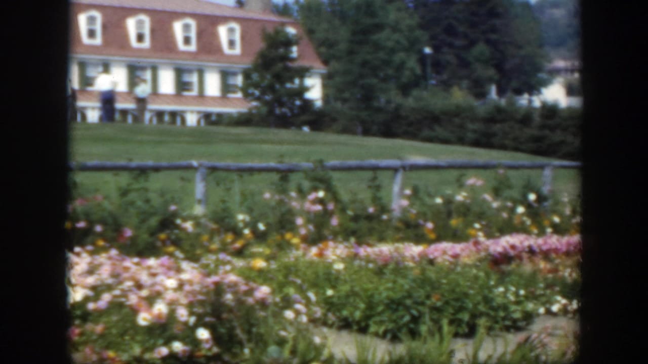 Flower beds and lawn in front of a white house with a red roof.