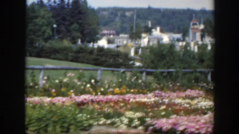 1950 Montreal frame showing landscaped flowers, lawn, fence, distant buildings, trees, and a small tower-like roofline at right.