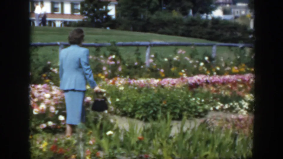 1950s Montreal home-movie frame of a woman in a blue suit walking away through a garden path lined with flowers.