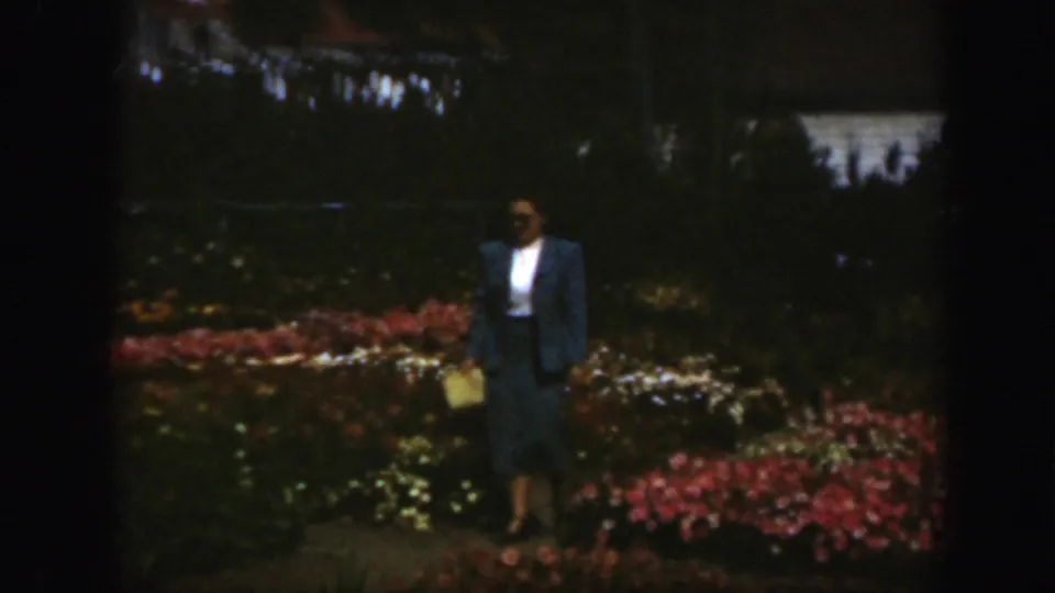 A woman in a dark skirt and blue jacket stands in shade among flower beds, holding a light-colored handbag in a Montreal garden.