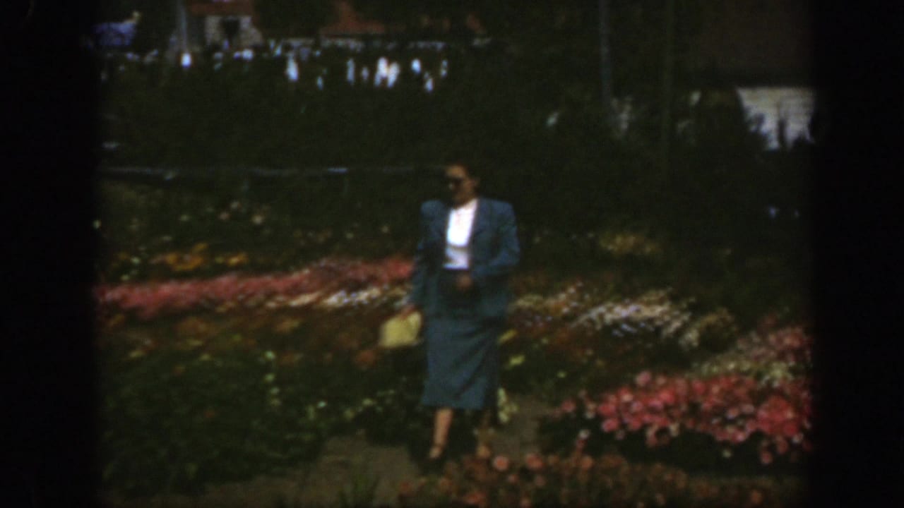 Woman in a blue suit walks beside colorful flower beds carrying a light purse.