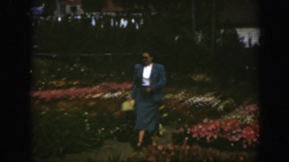 In 1950 Montreal home-movie footage, a woman in sunglasses and a blue suit walks along a garden path with flower beds behind her.
