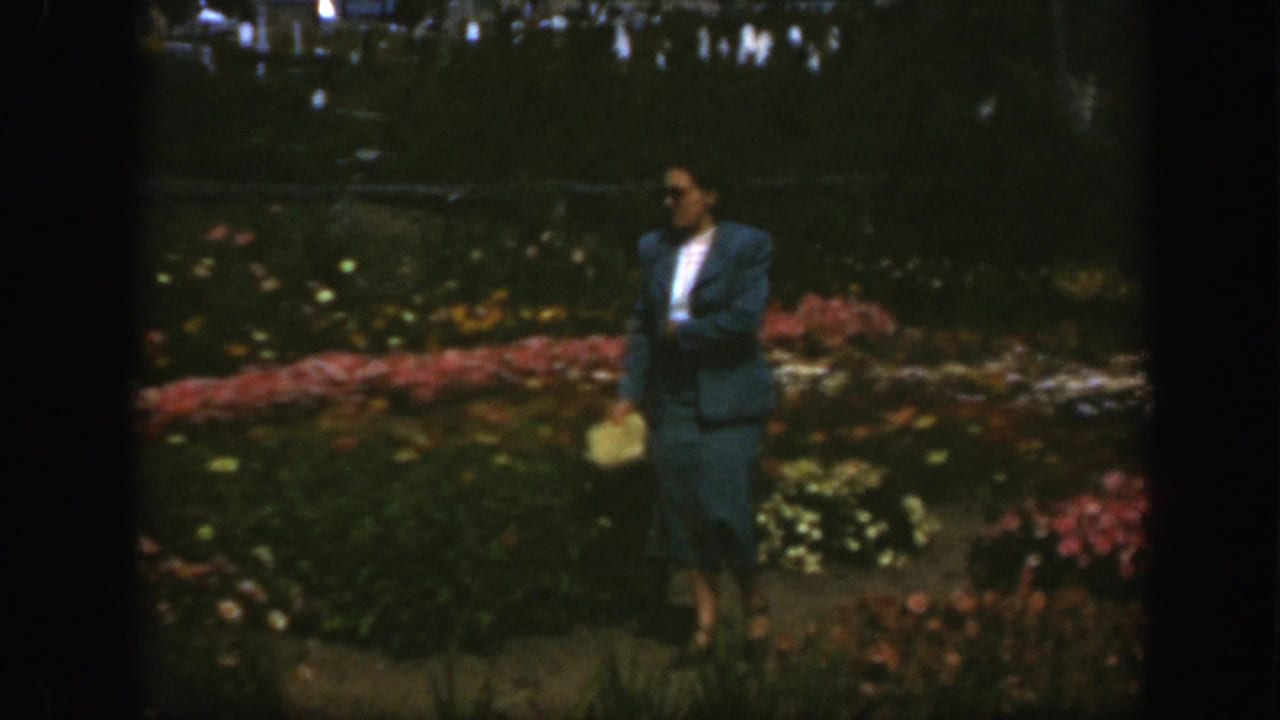 Woman stands or walks near flower beds with other garden visitors in the distance.