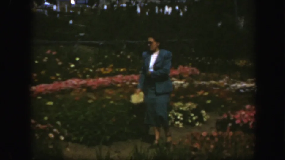 The woman in a blue skirt suit and sunglasses moves through a Montreal garden, with rows of flowers and distant visitors visible.