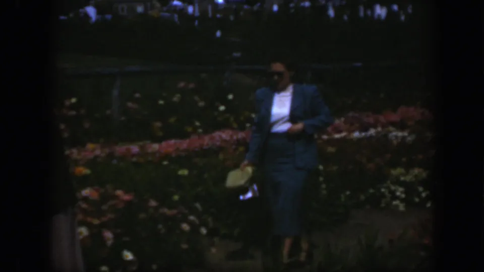 A woman in mid-century dress approaches the camera through a flower garden in 1950 Montreal, holding a light purse at her side.