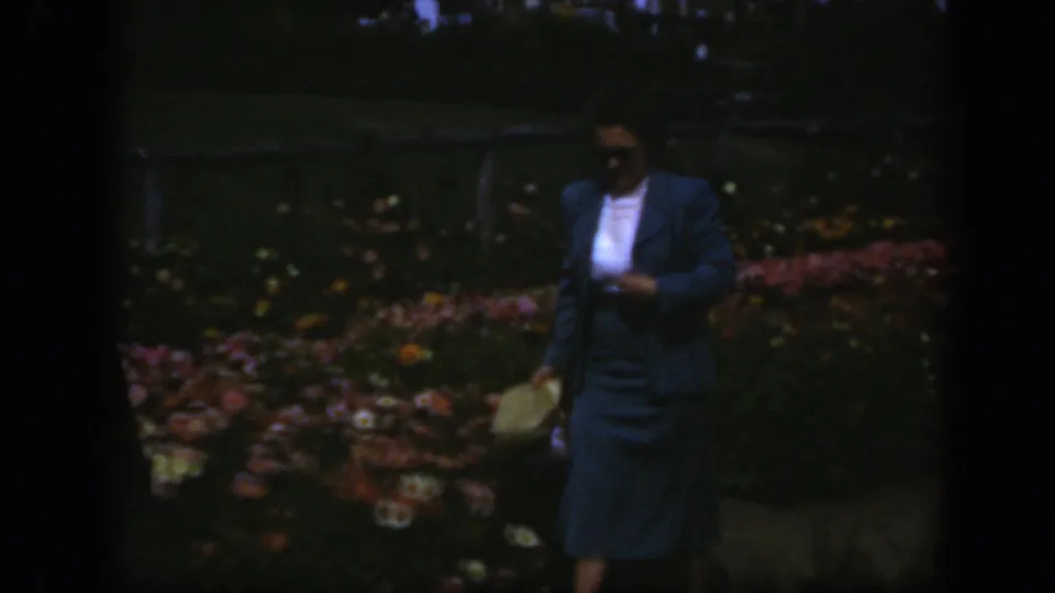 Near the end of the 8mm clip, the woman passes closer to camera in a blue suit, with garden flowers and dark film edges visible.