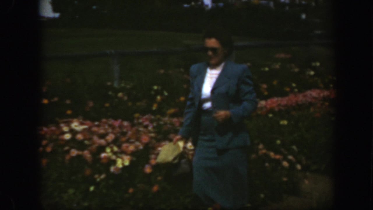 Woman in sunglasses and a blue suit walking beside flower beds.