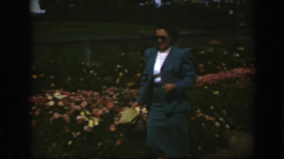In 1950 Montreal home-movie footage, a woman in sunglasses and a blue suit walks through a garden holding a small handbag.