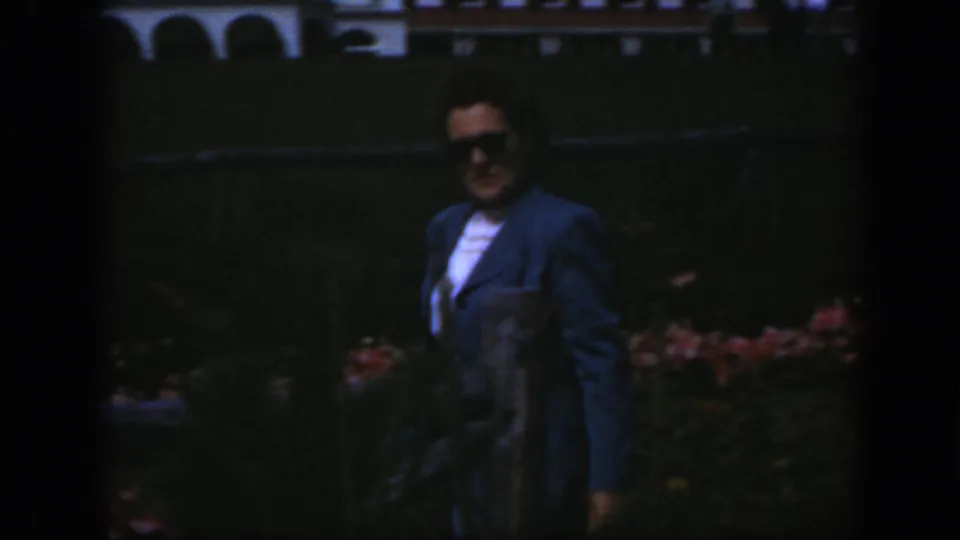 A woman in sunglasses and a blue suit appears to smile toward the camera while standing near garden flowers.