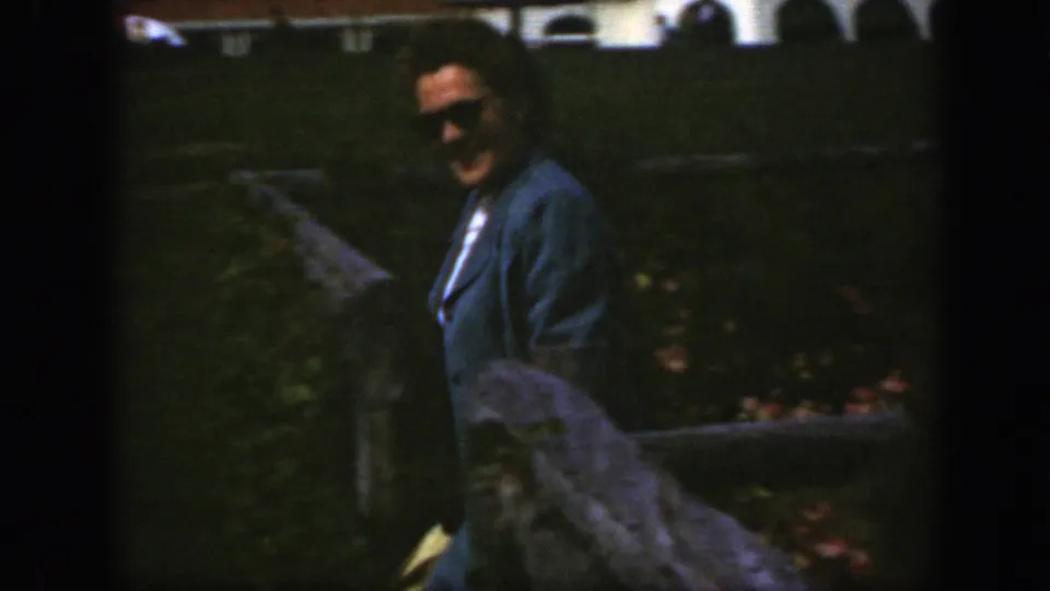 The woman stands near a garden path or stonework, with flower beds and lawn visible behind her in the 1950 Montreal footage.