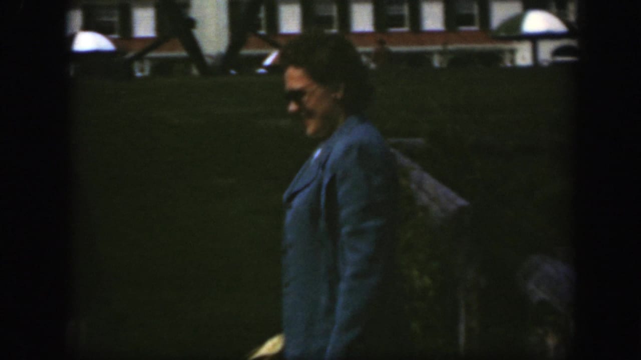 Woman in a blue coat walks across a lawn with a brick building behind her.