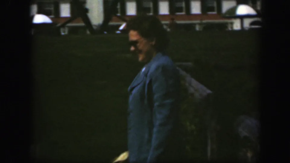In 1950 Montreal footage, a woman in a blue coat and sunglasses walks on grass near a large brick building.