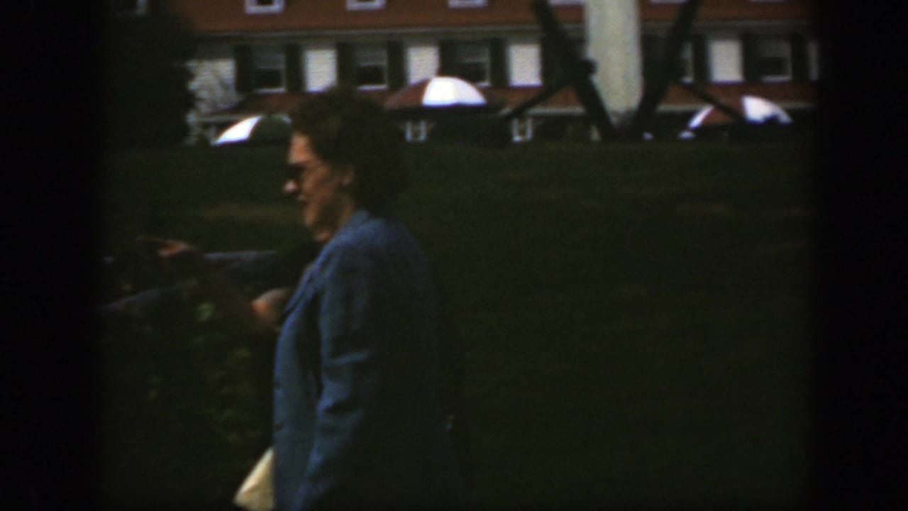 Woman in sunglasses walks in profile on a lawn beside a low rail.