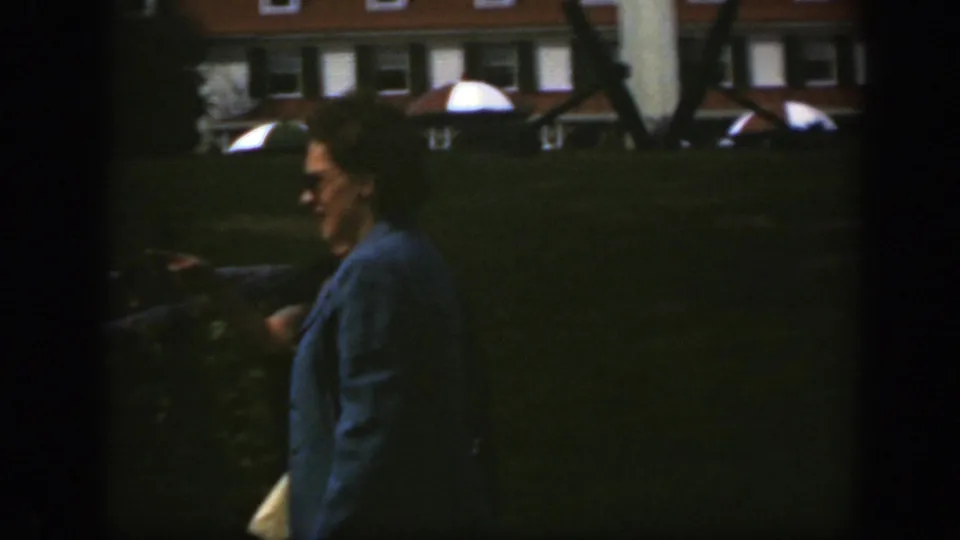 The woman continues walking in profile across a grassy Montreal lawn, with a brick building in the background.