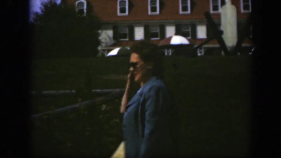 In the 1950 Montreal home movie, the woman pauses or turns slightly with one hand near her face and a pale bag at her side.