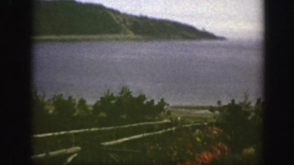 1950s 8mm landscape near Montreal with calm blue water, a wooded hillside, foreground shrubs, and a rail fence.