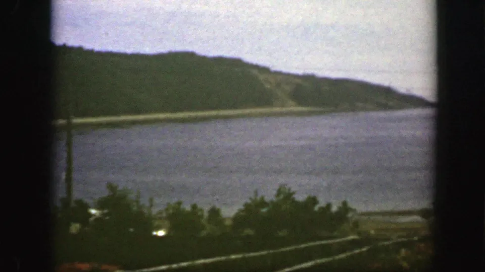 1950s 8mm view near Montreal looking across blue water toward a tree-covered hillside, with shrubs and fence rails below.