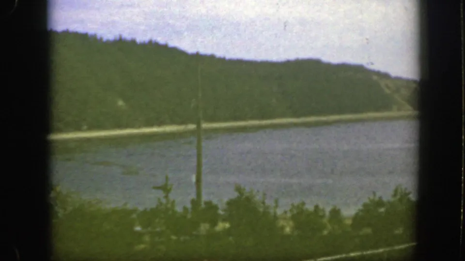 1950s 8mm Montreal-area waterfront view with a green hillside, calm water, foreground shrubs, fence rails, and a utility pole.