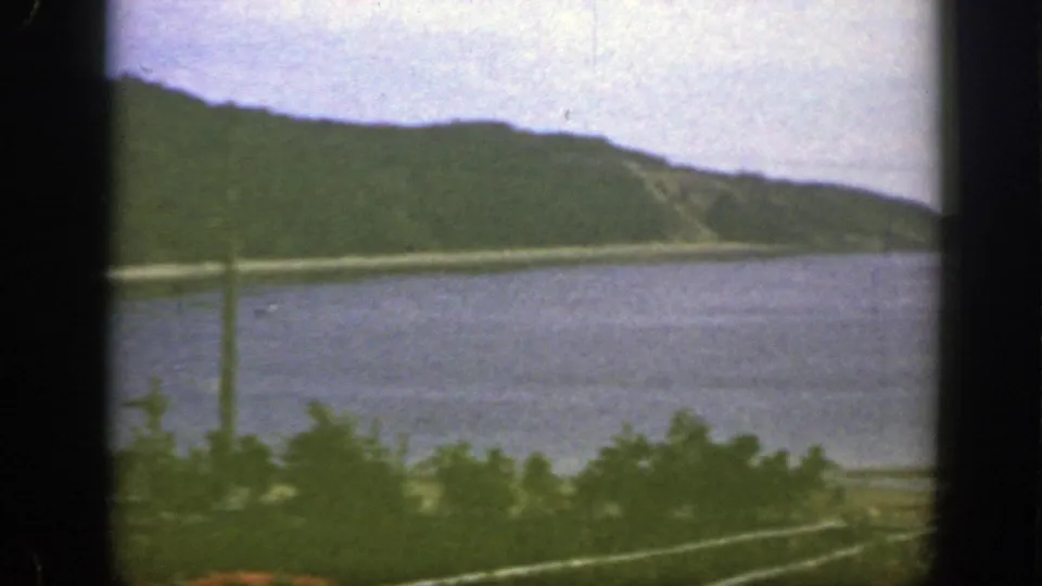 1950 8mm home-movie view near Montreal showing calm water, a green hillside, foreground vegetation, and a utility pole.