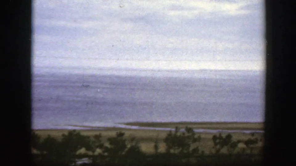 1950 8mm Montreal-area view across calm water toward a low shoreline, with trees below and dark frame edges suggesting a window view.
