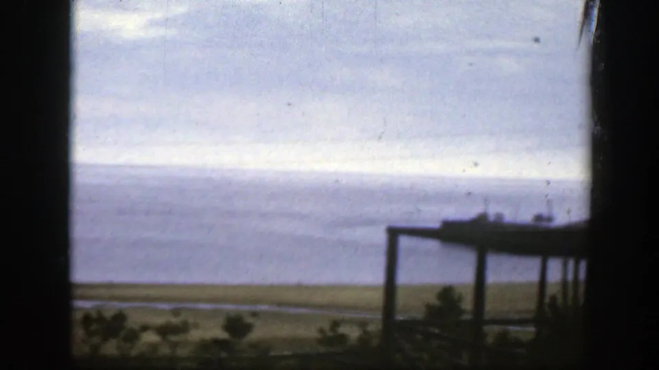 1950 8mm Montreal-area waterfront view with cloudy sky, calm water, low shore, foreground vegetation, and a partial structure at the right edge.