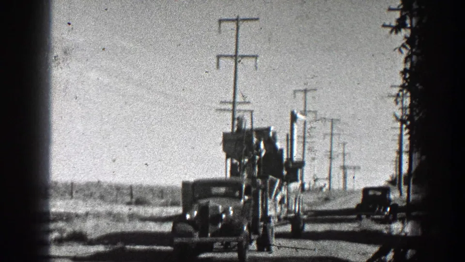 1936 California 8mm frame showing a vehicle or tractor-like hauler pulling a large wheeled apparatus along a dusty rural road lined with utility poles.