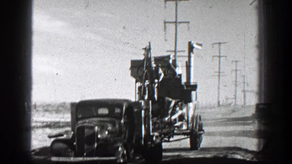 1936 California 8mm frame showing a close roadside view of older motor traffic and a large towed apparatus beneath a line of utility poles.