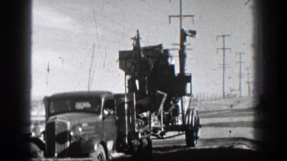 1936 California home-movie frame with an older vehicle beside a tall towed equipment rig, set against a dusty rural road and repeated utility poles.