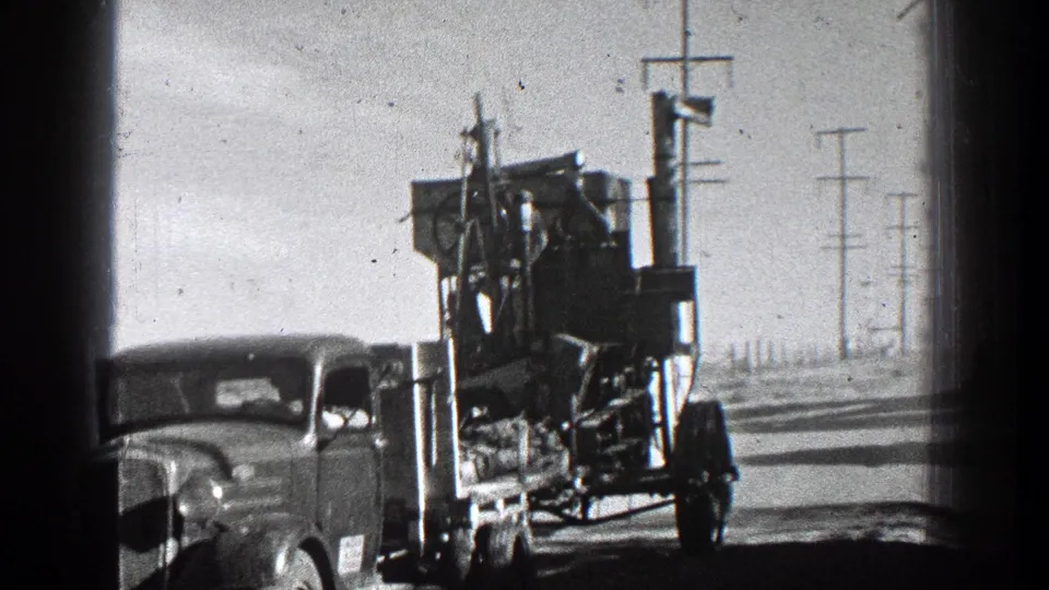 1936 California home-movie frame showing a vintage truck towing farm machinery past roadside utility poles in bright daylight.
