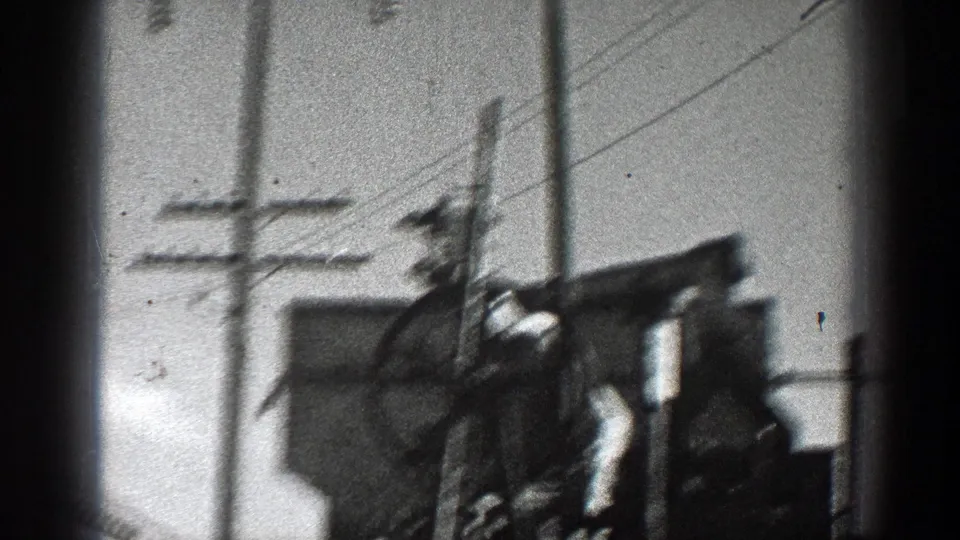 Black-and-white 8mm frame from California showing pole-mounted metal utility equipment and overhead lines in soft focus.