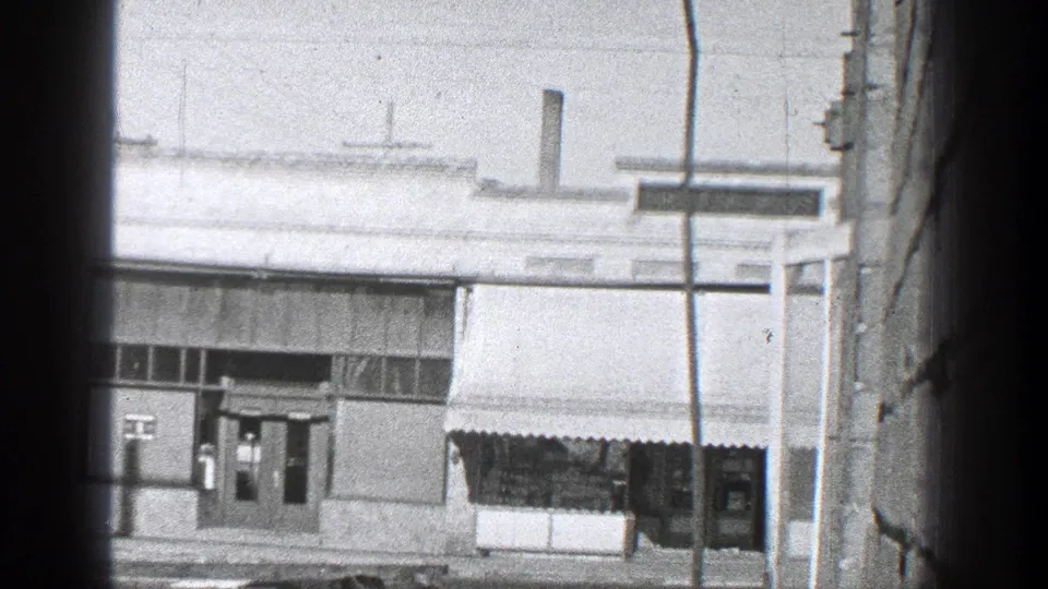 Black-and-white 8mm view of a California storefront, awnings, doors, and utility pole from a street-side vantage.