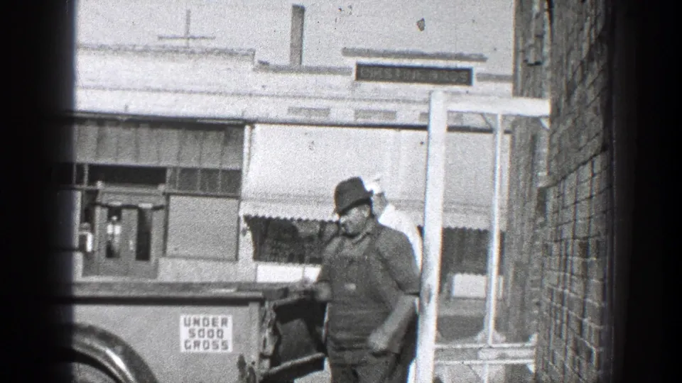 The worker remains beside the rear of the truck, with a storefront awning and a visible sign panel in the 1936 California street scene.