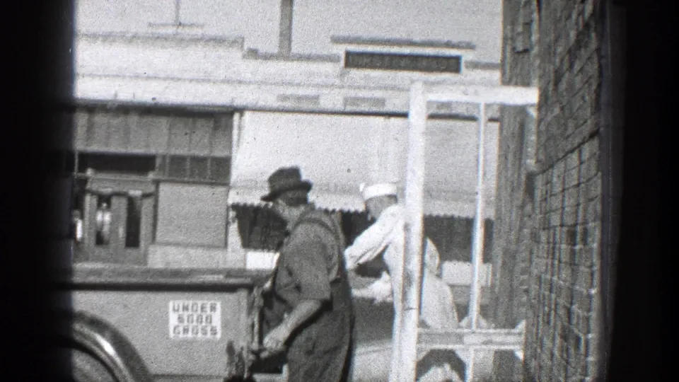A worker in overalls passes the truck rear while a second man in a white uniform stands near the storefront in California archival footage.